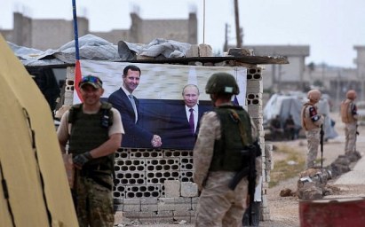 Members of the Russian military stand past a banner showing Russian President Vladimir Putin, right, shaking hands with Syrian President Bashar Assad, at Abu al-Zuhur checkpoint in the western countryside of Idlib province on June 1, 2018. (AFP PHOTO / George OURFALIAN)